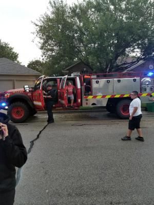 Fire truck parked on street, firefighter and man standing by open door, another man walks by.