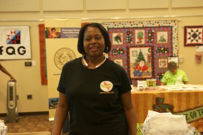 Woman in black shirt smiles at a booth, a quilt and sign in the background.
