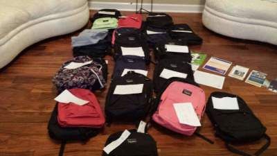 Backpacks of various colors with white labels, on a wooden floor, near a couch.