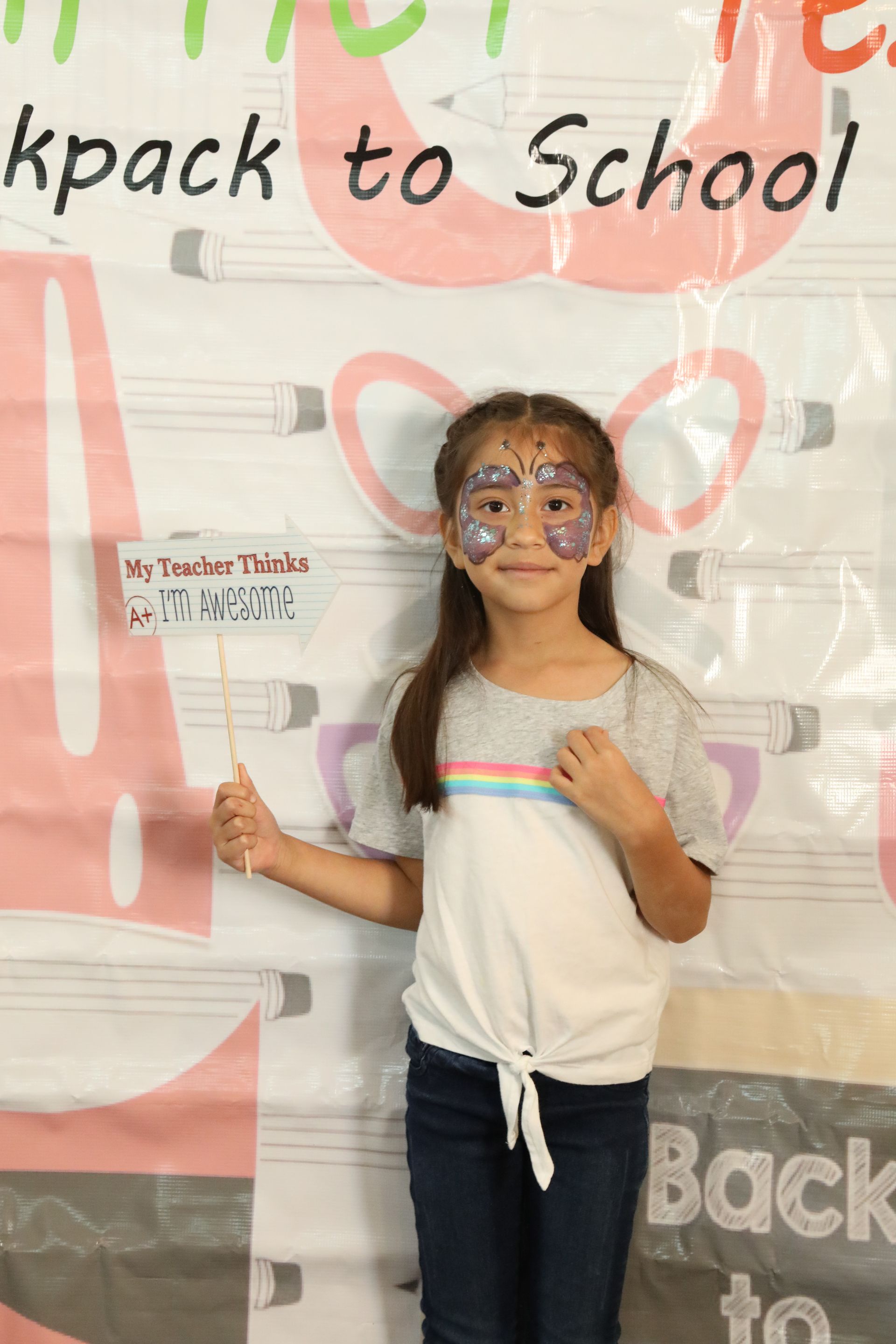 Girl with glitter face paint poses for a back-to-school photo, holding a sign.