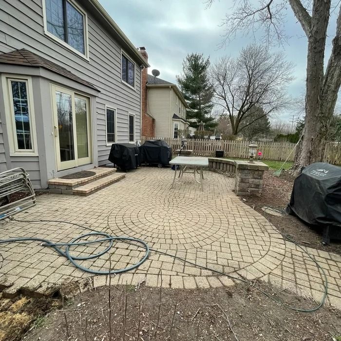 Backyard stone patio beside a house, with grill covers, table, hose, and trees in the yard