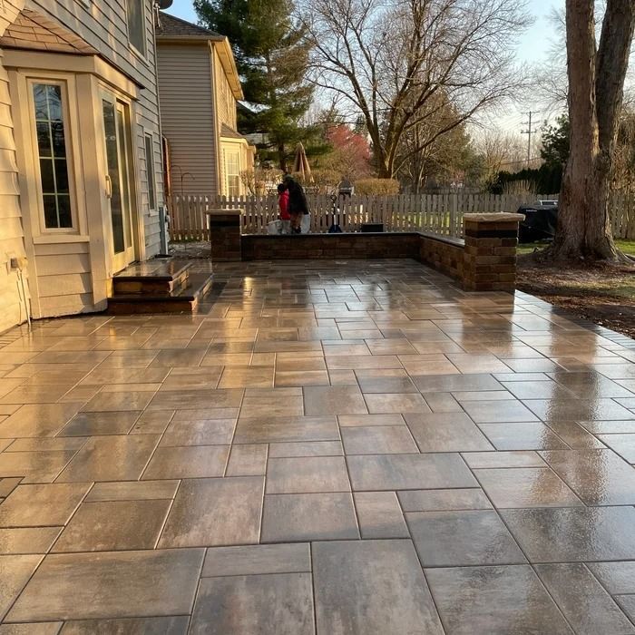 Stone patio beside a house, reflecting warm sunlight, with trees and a person in the background.