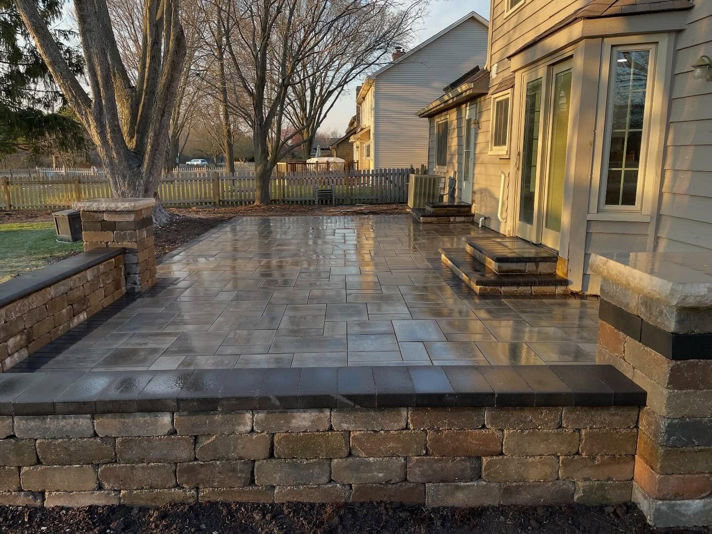 Stone patio beside a beige house at sunset, with brick planters and leafless trees in the yard