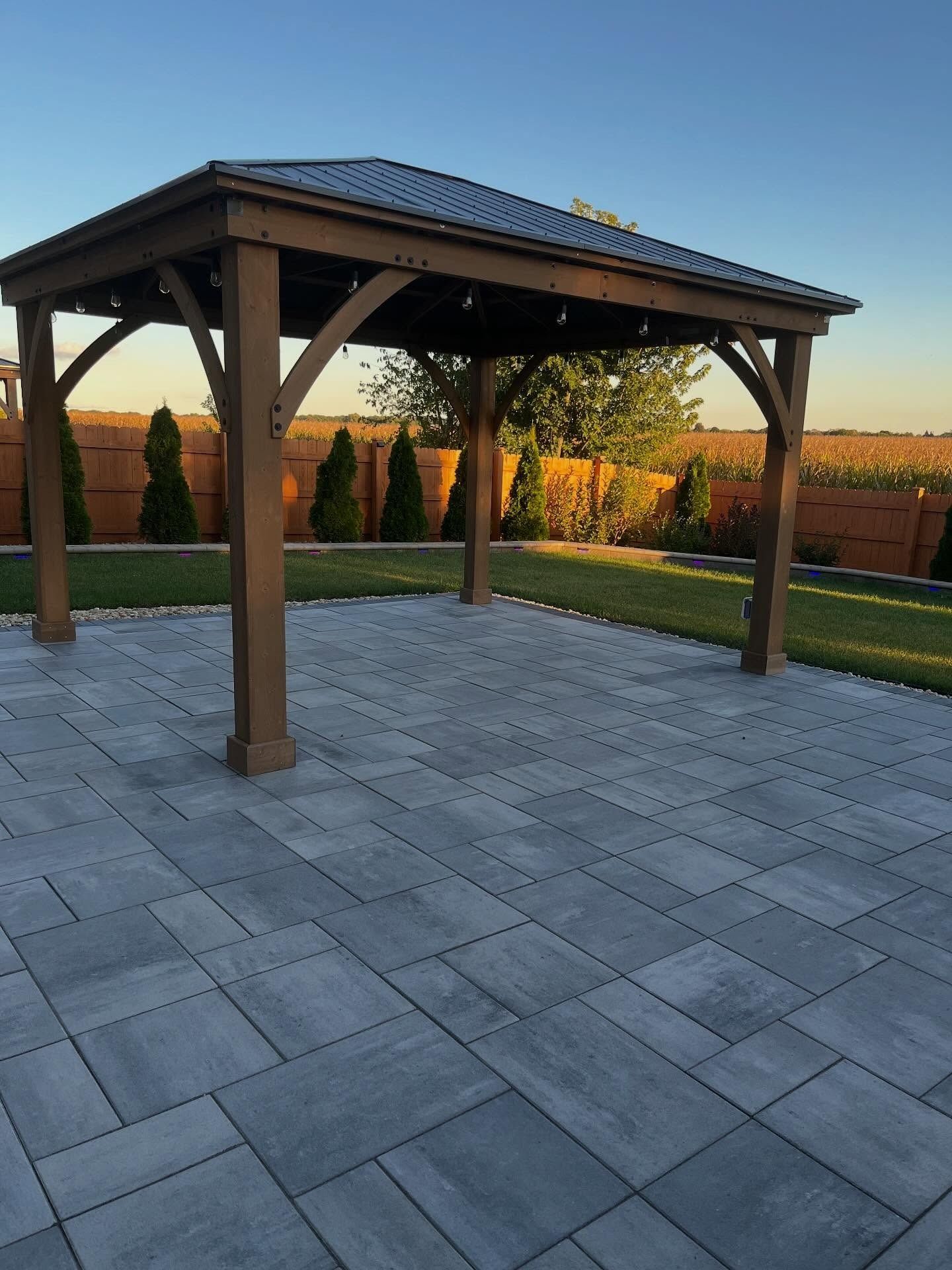Covered patio pavilion with wooden posts on a gray stone patio in a landscaped backyard at sunset