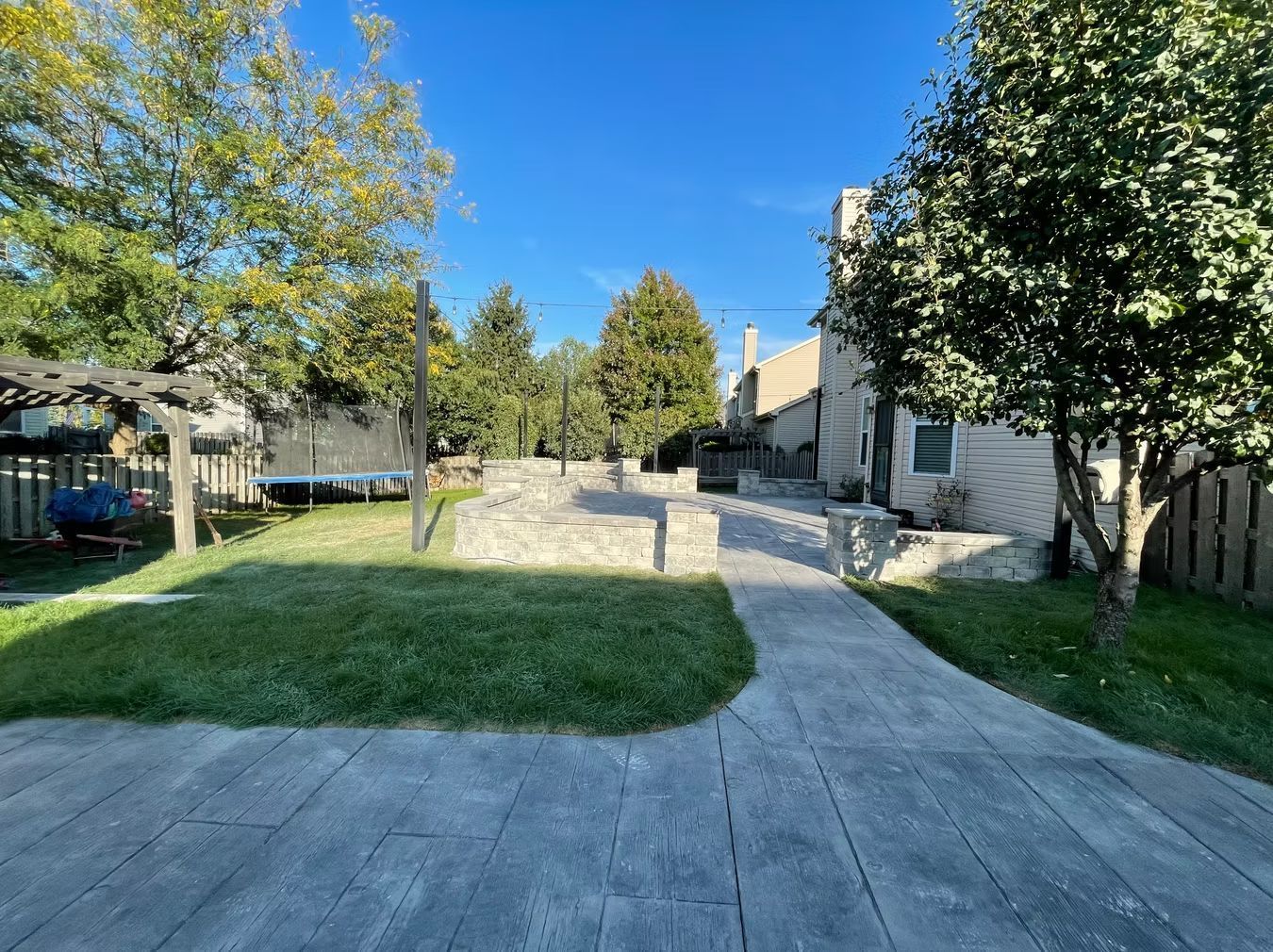 Sunny paved walkway through a landscaped courtyard with trees, grass, and stone buildings.