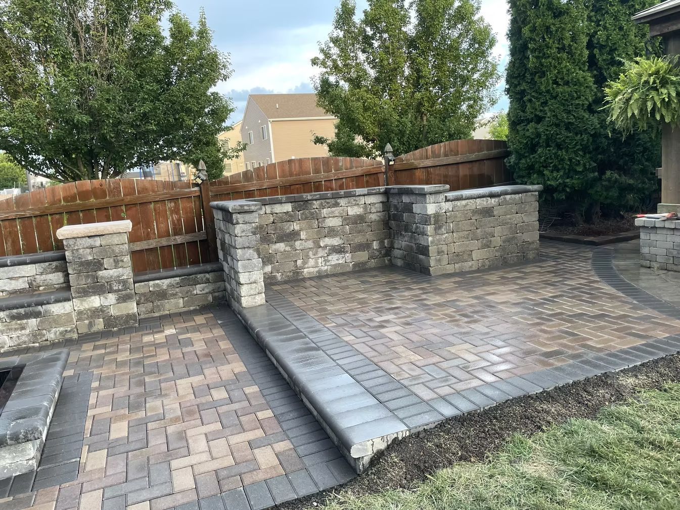 Paver patio with stone retaining walls, wooden fence, and trees in a backyard.