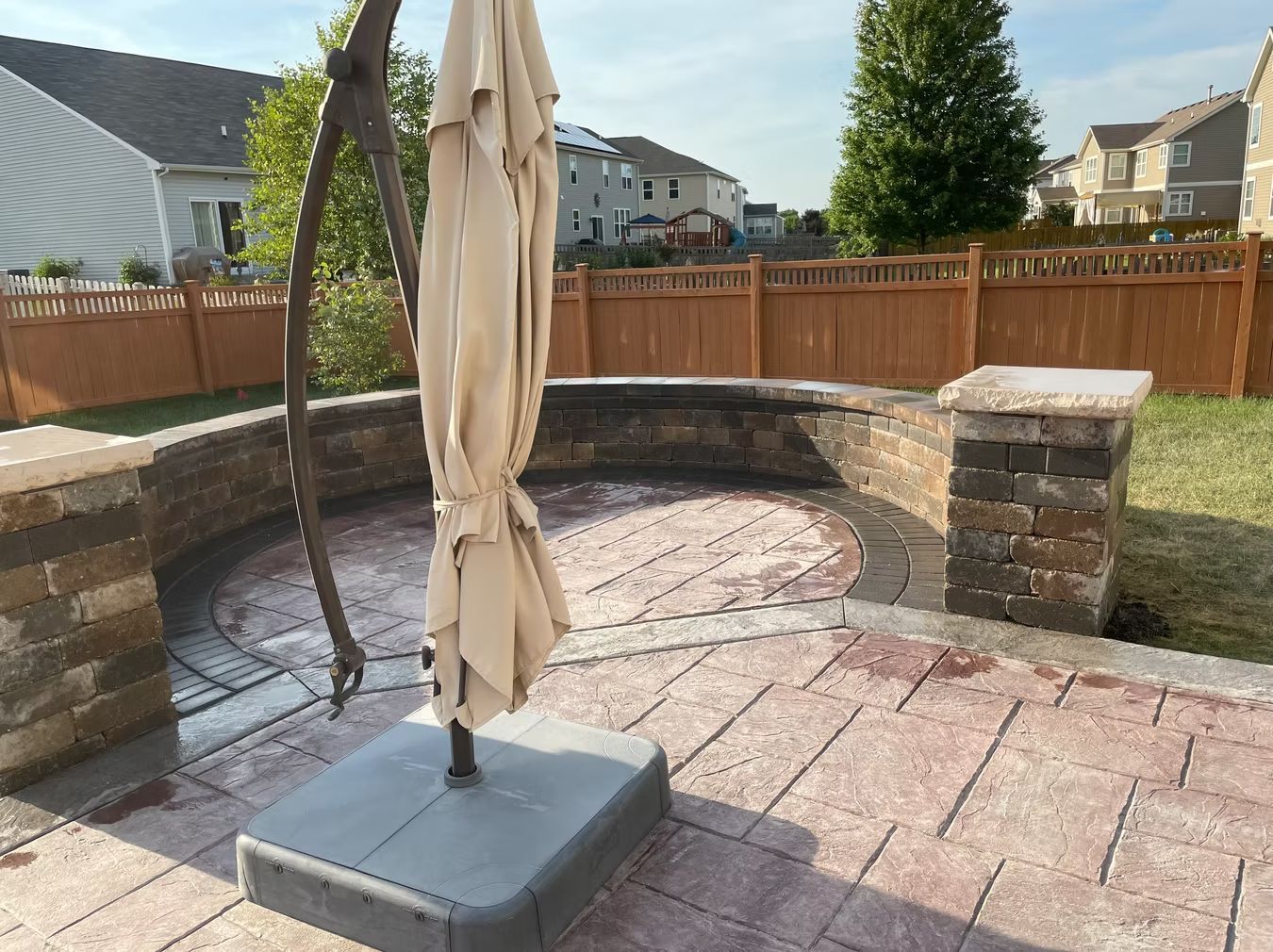 Backyard patio with a folded beige umbrella, brick fire pit, and wooden fence in the background