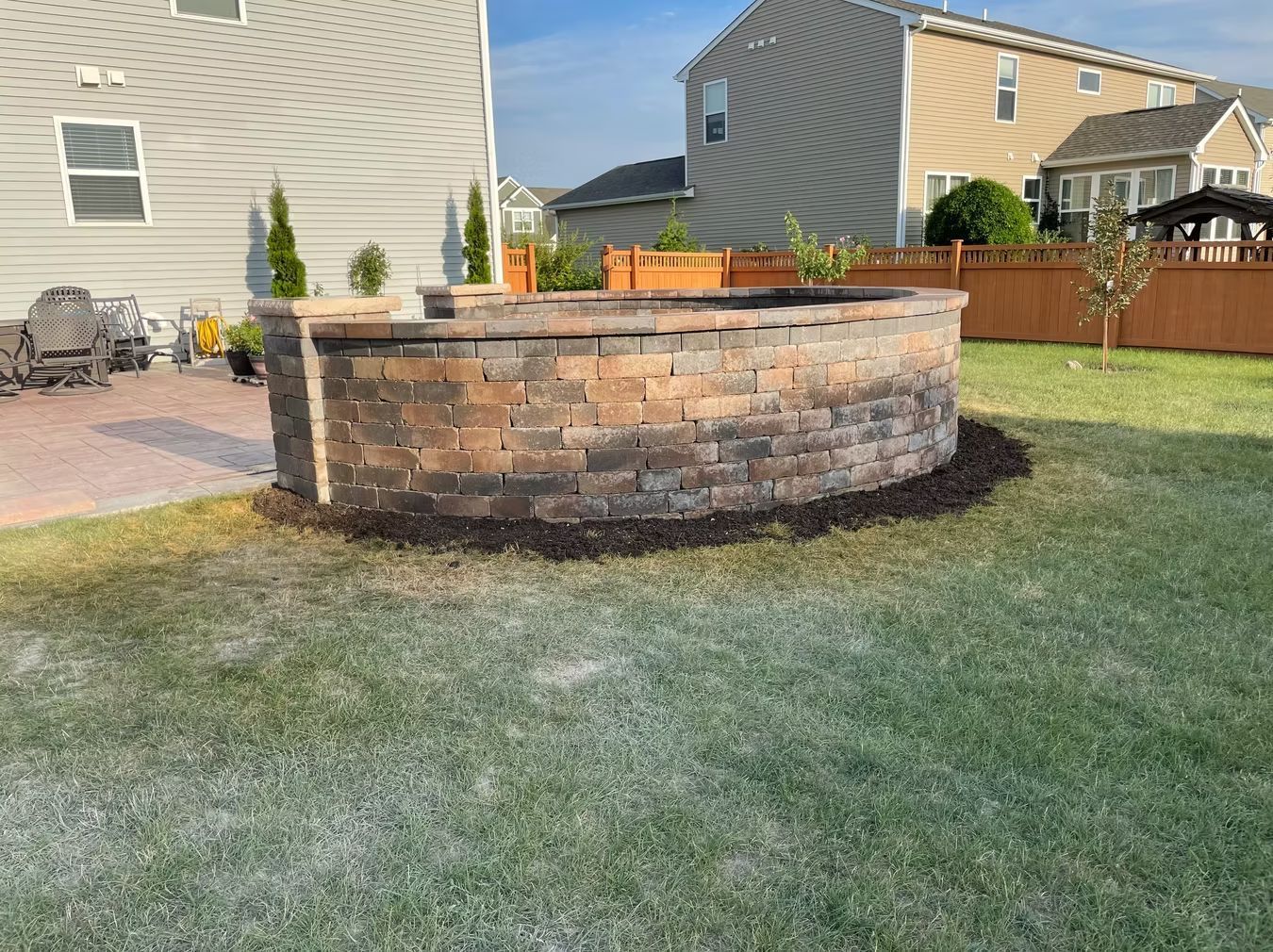 Backyard brick fire pit with wood burning, surrounded by grass and a patio near suburban houses