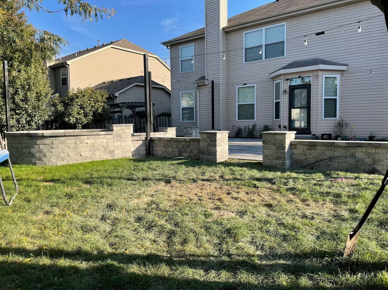 Backyard with stone retaining wall, grass lawn, and a two-story house under clear blue sky