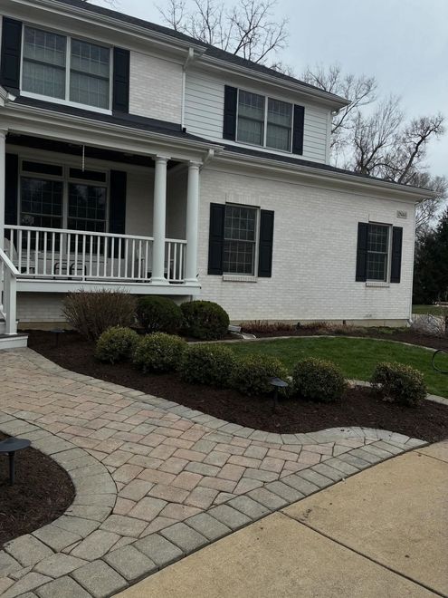 White two-story house with black shutters, front porch, and brick walkway through trimmed shrubs