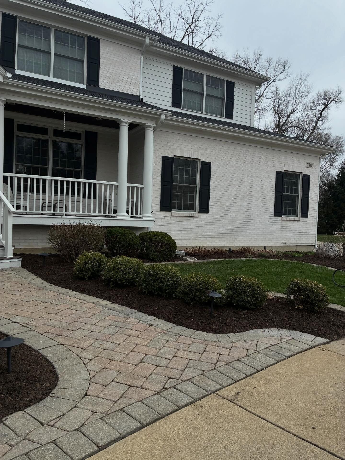White two-story house with black shutters, front porch, and brick walkway in a landscaped yard