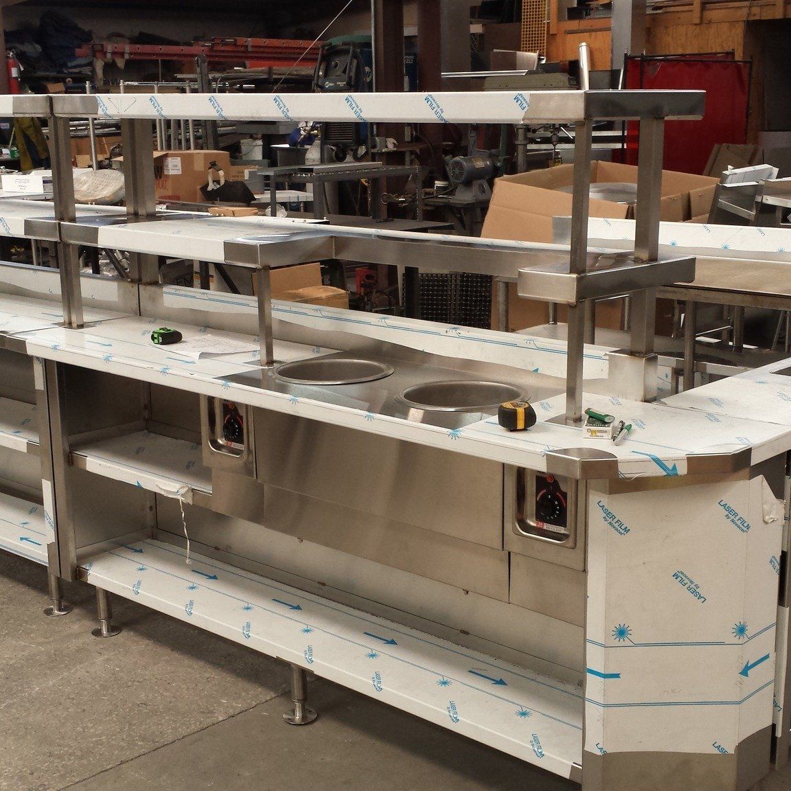 Large stainless steel food service counter/display unit in a workshop, partially wrapped in protective film.