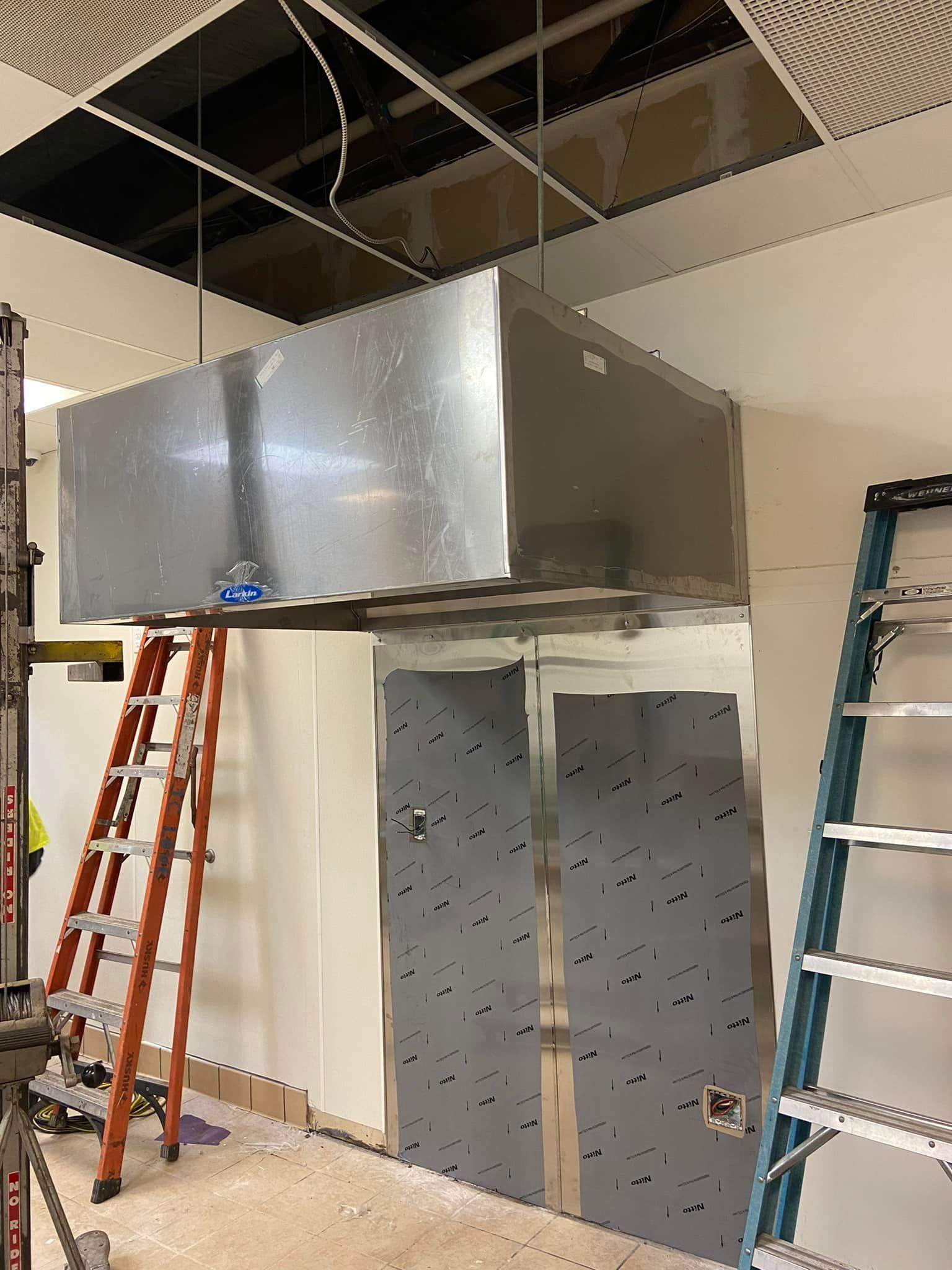 Ladders beside a large silver HVAC duct in a room under construction with unfinished walls and exposed ceiling