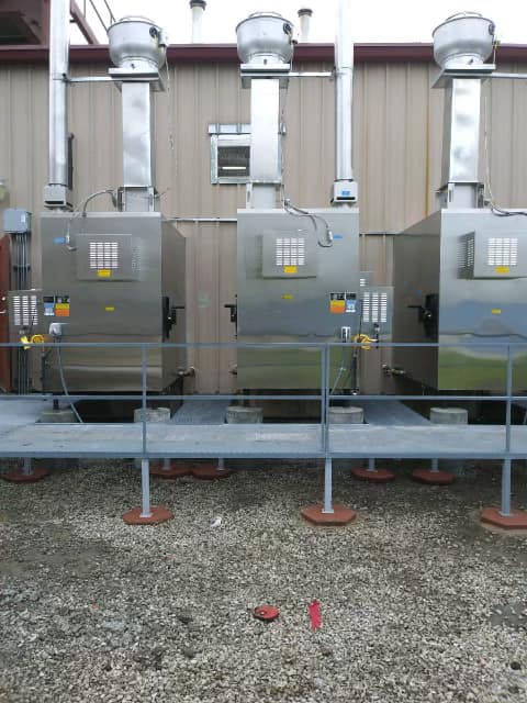 Three gray industrial air units on a metal platform outside a building, with ductwork above.