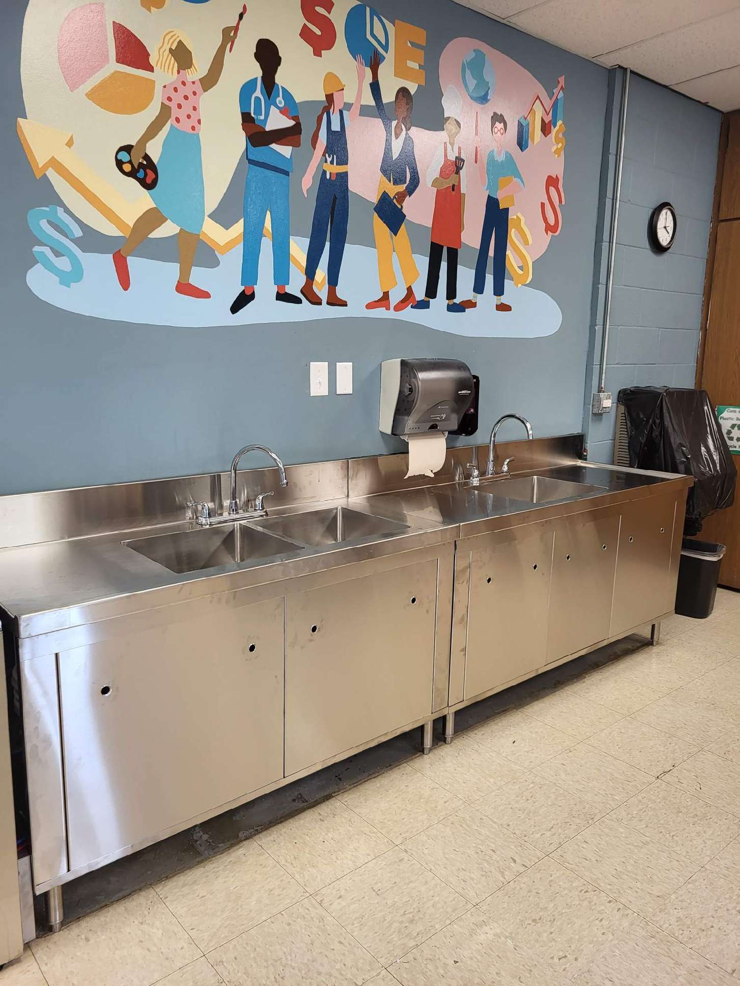 School cafeteria drink station with stainless steel sinks, mural, and a trash can on a tiled floor