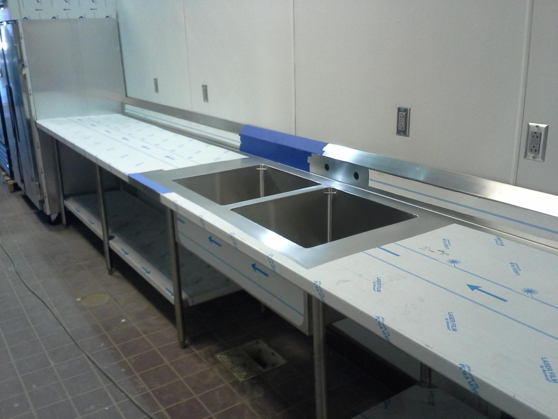 Stainless steel commercial kitchen counter with sink basins and a white work surface along a wall