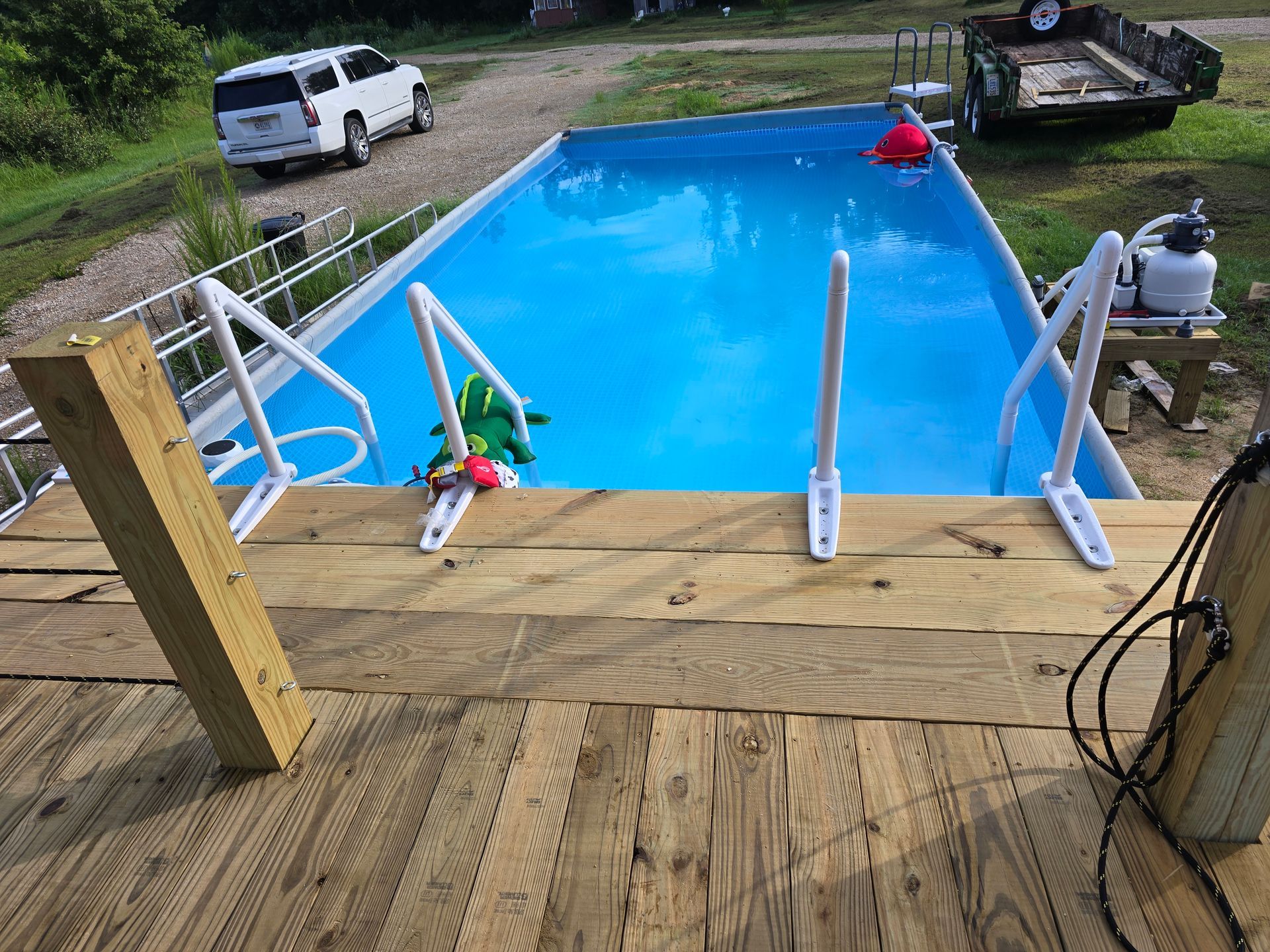 A rectangular above-ground pool with white ladders sits next to a wooden deck, a truck and trailer in background.
