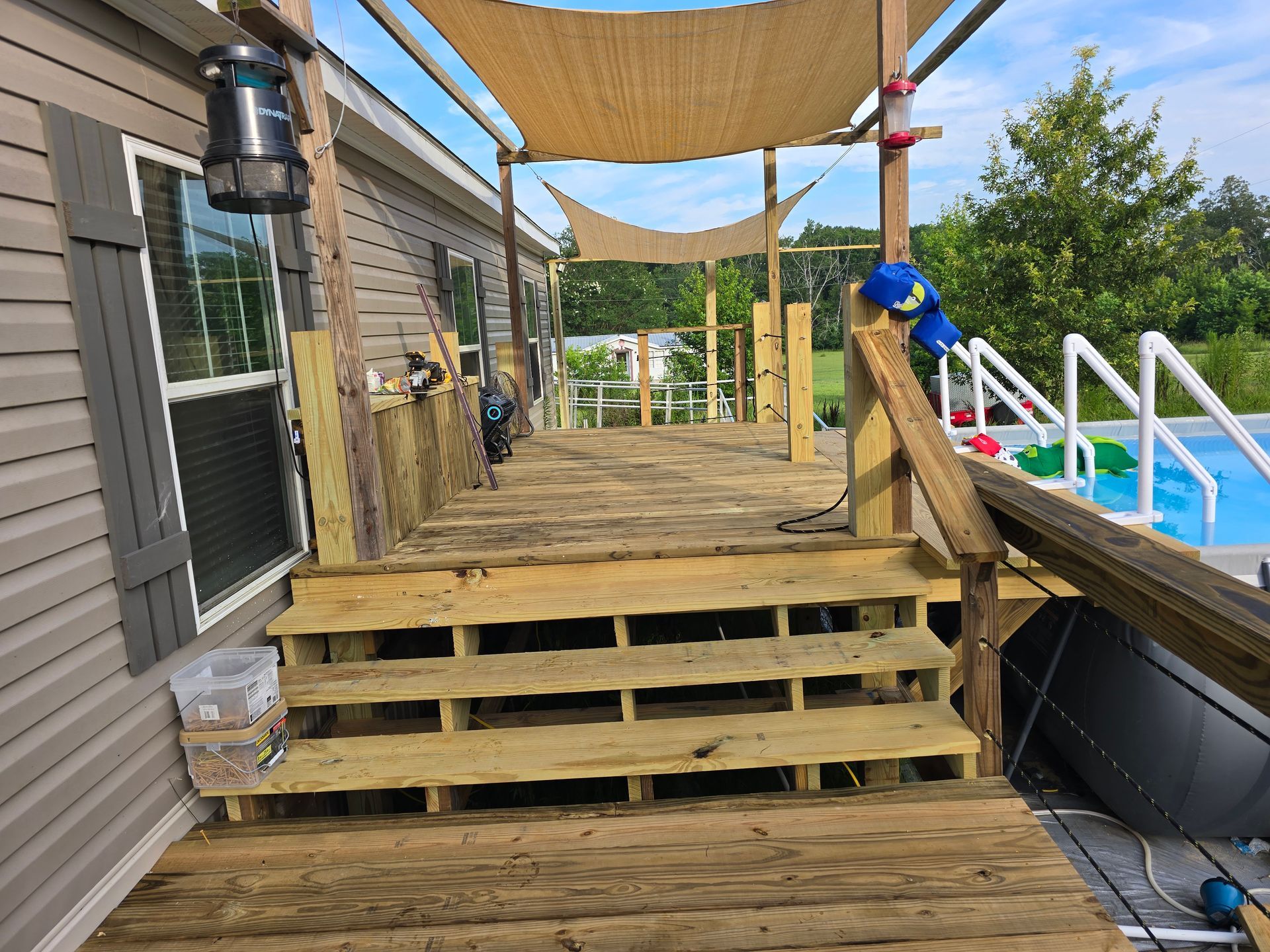 Wooden deck with steps leading to a pool area; a tan shade sail covers part of the deck.