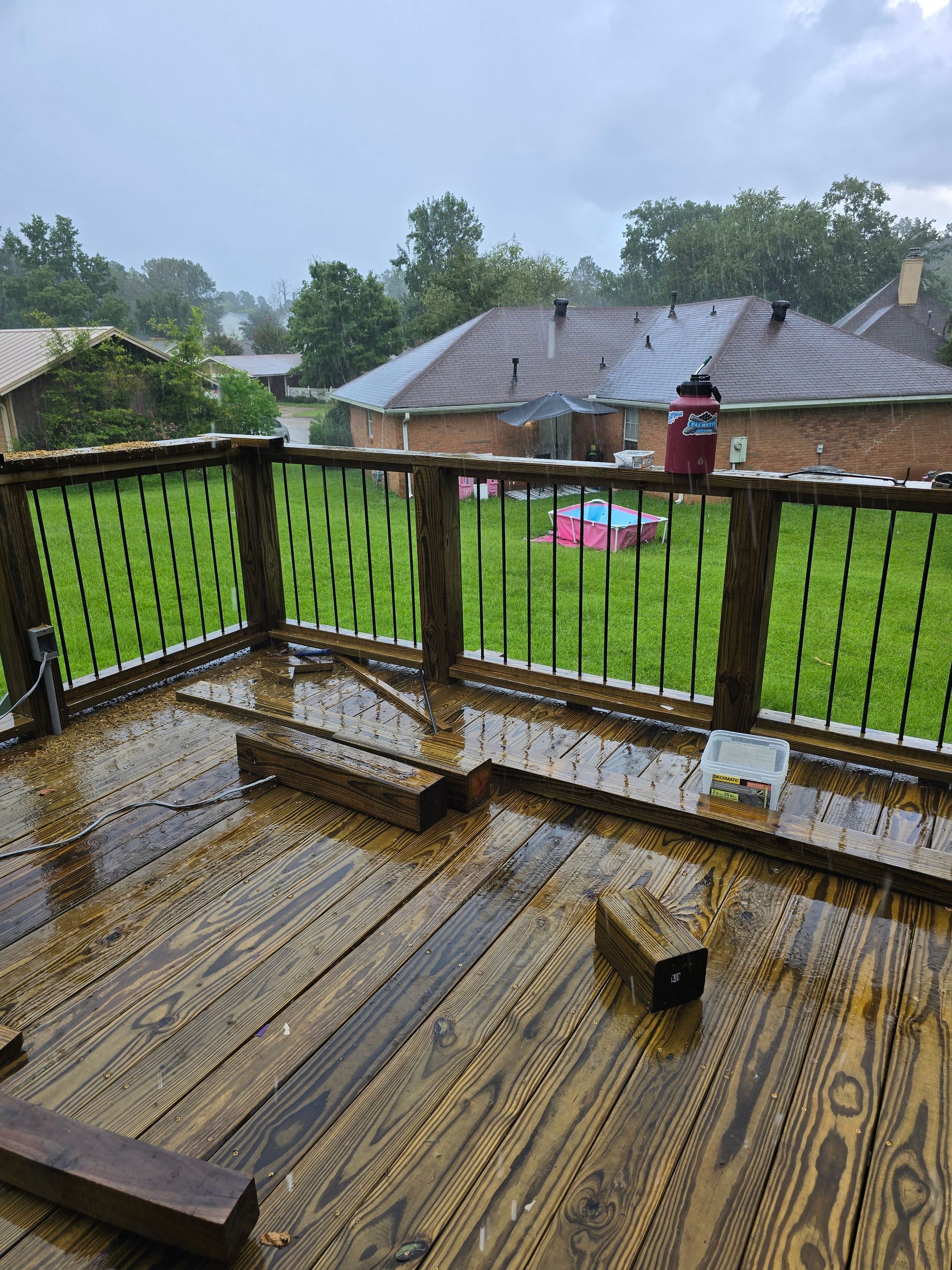 Rainy wooden deck with black railing overlooking a green yard and brick houses.