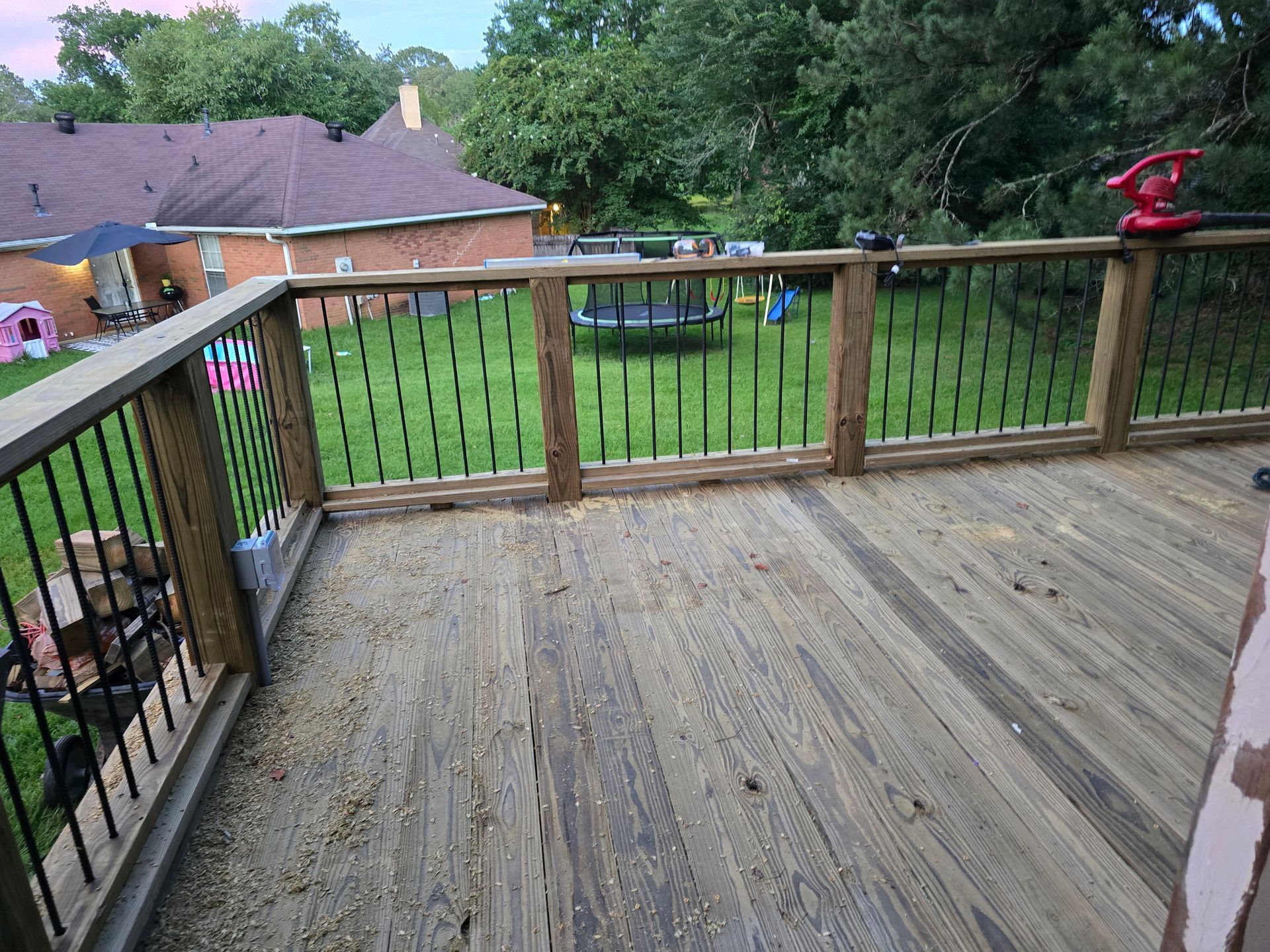 Wooden deck with black railings overlooking a grassy backyard with a trampoline and a house in the background.