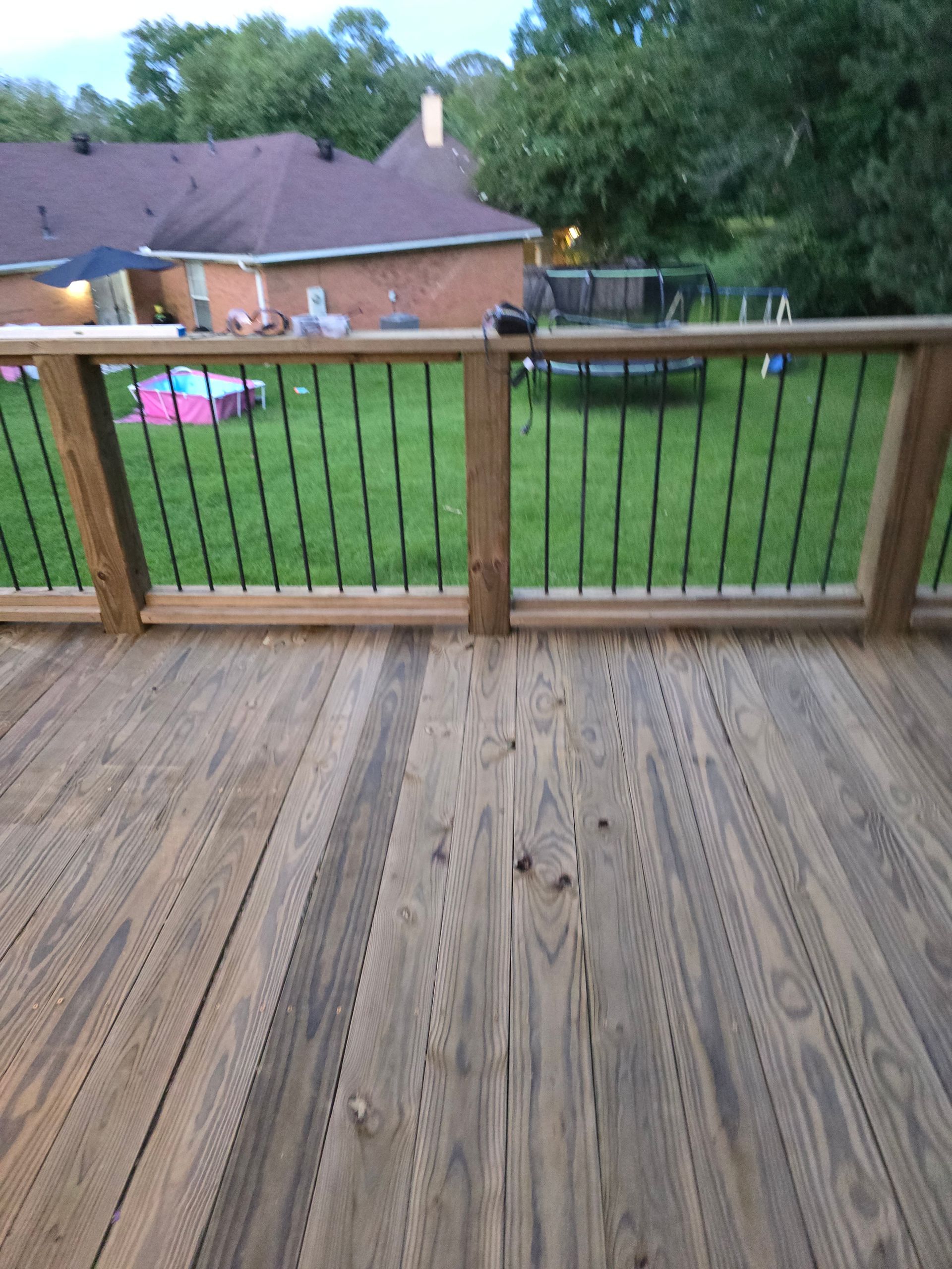 Wooden deck overlooking a grassy backyard with a house in the background. Black railings, wooden posts.