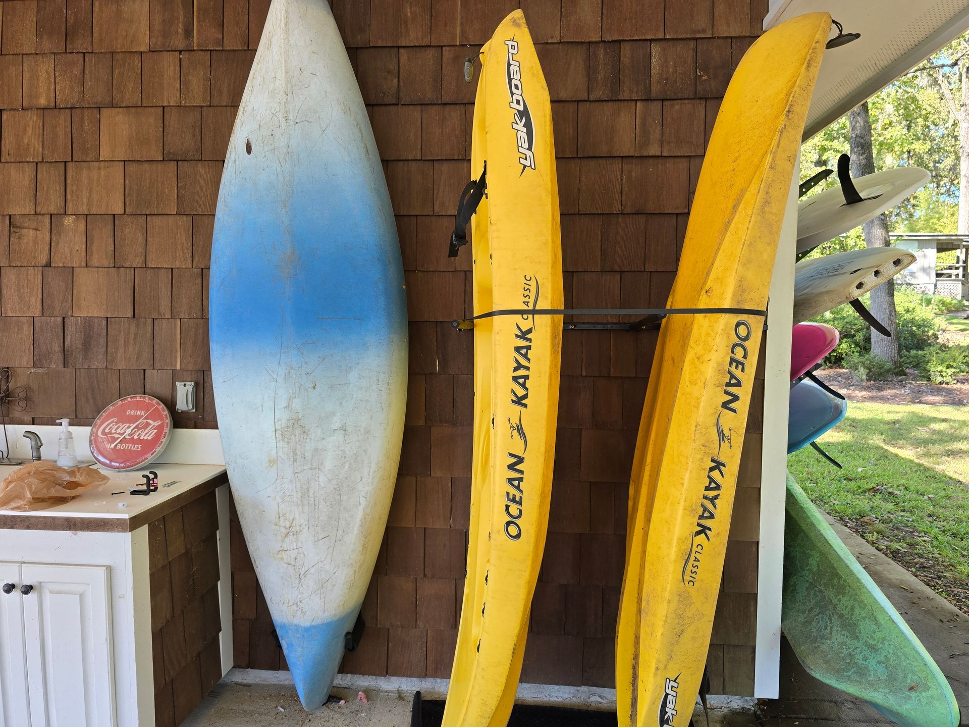 Surfboards and kayaks lean against a brown shingle wall, next to a white cabinet and counter.