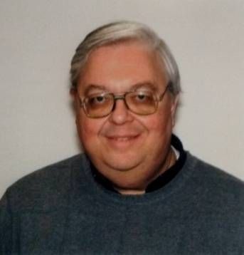 Smiling person with glasses and gray hair wearing a dark sweater against a plain background