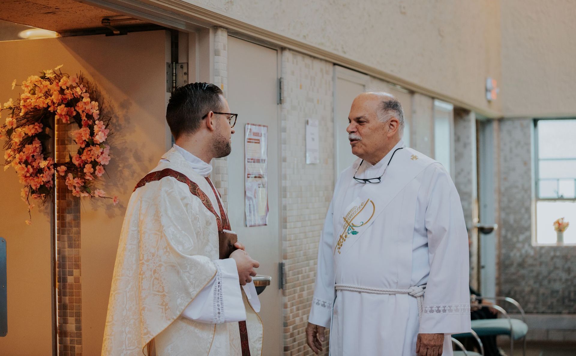 Two clergy members in white robes converse indoors near a decorated wall