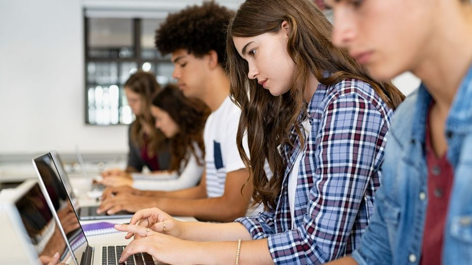 Students using laptops in a classroom, focused on typing at a long table.