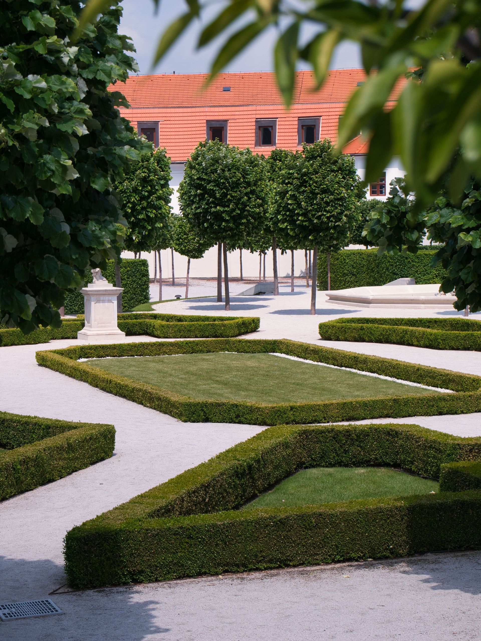 Formal garden with geometric hedges, gravel paths, and trimmed trees in front of a red-roofed building