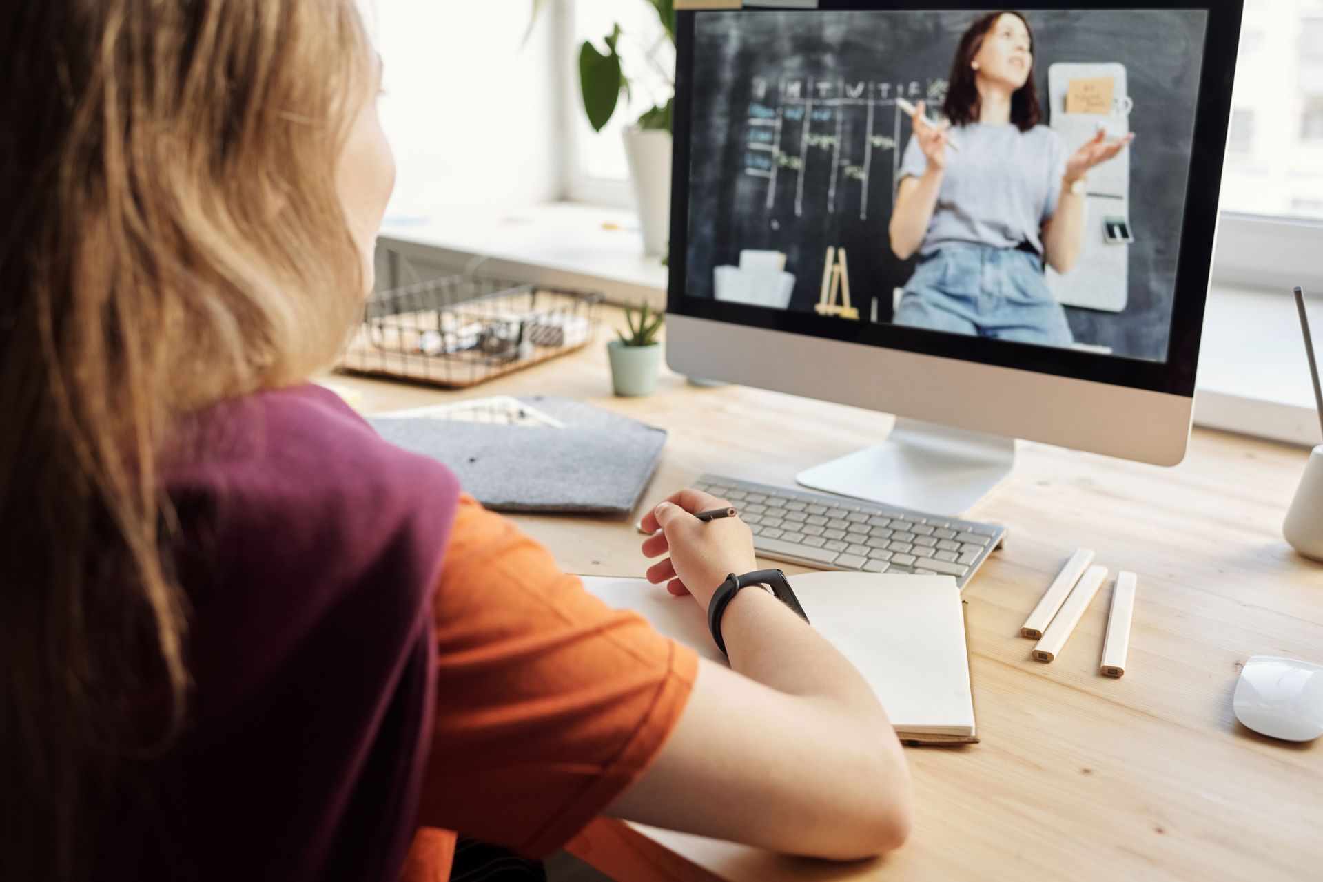 Person taking notes during a video call at a desk, watching a woman speaking on a computer screen