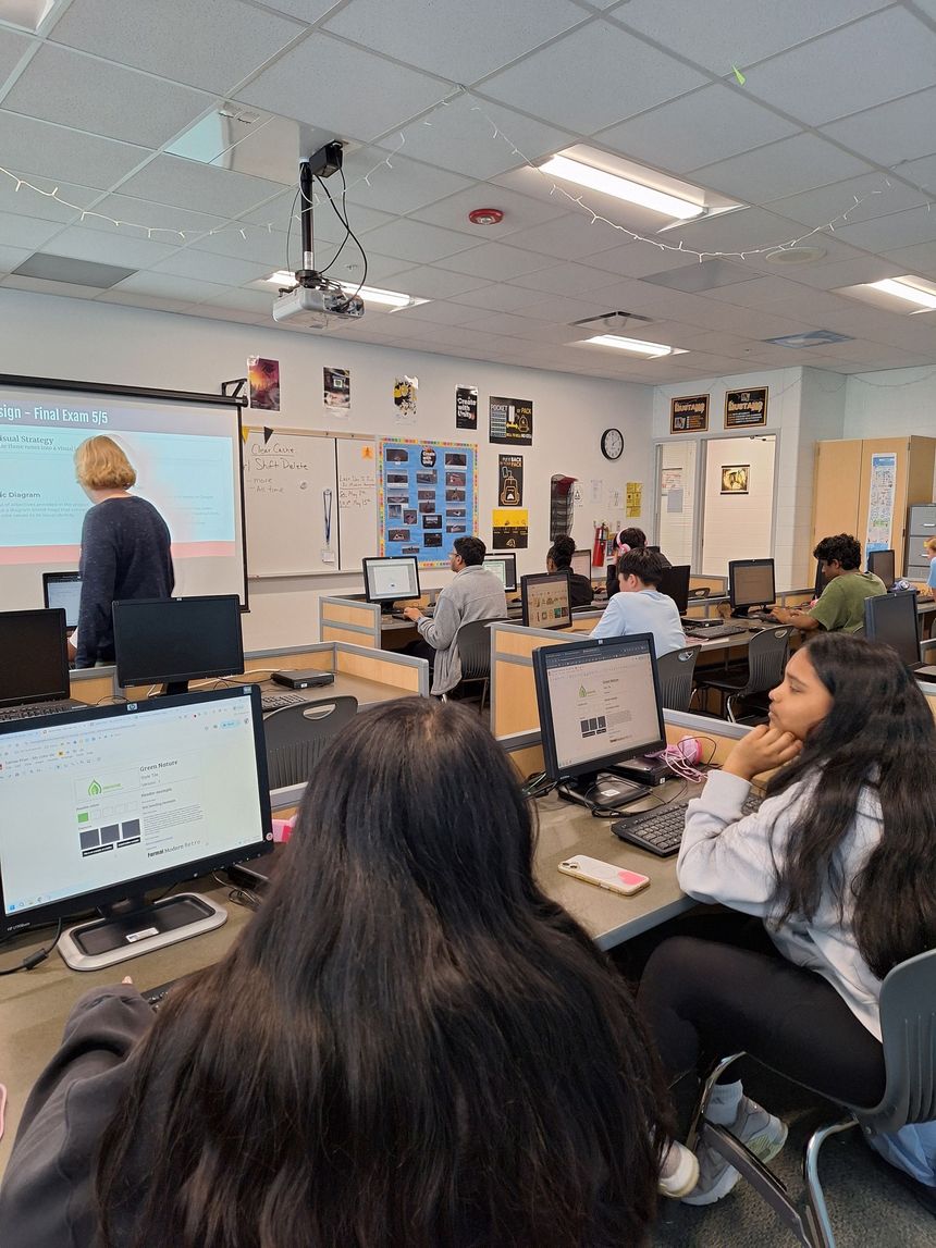 Computer lab classroom with students at desktop computers and a teacher at the front by a whiteboard.