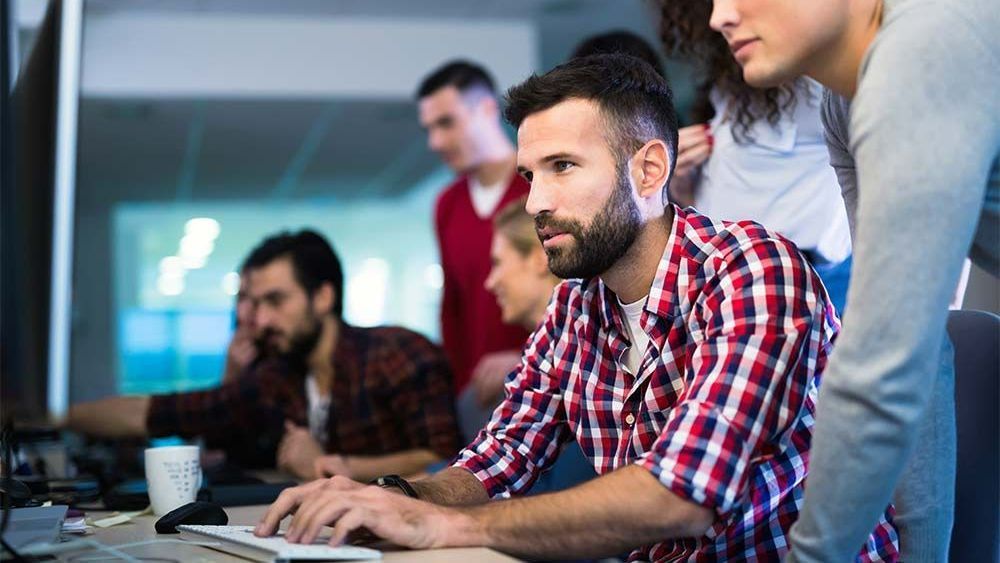 Team of coworkers collaborating around a laptop in an office, with one man typing in the foreground.