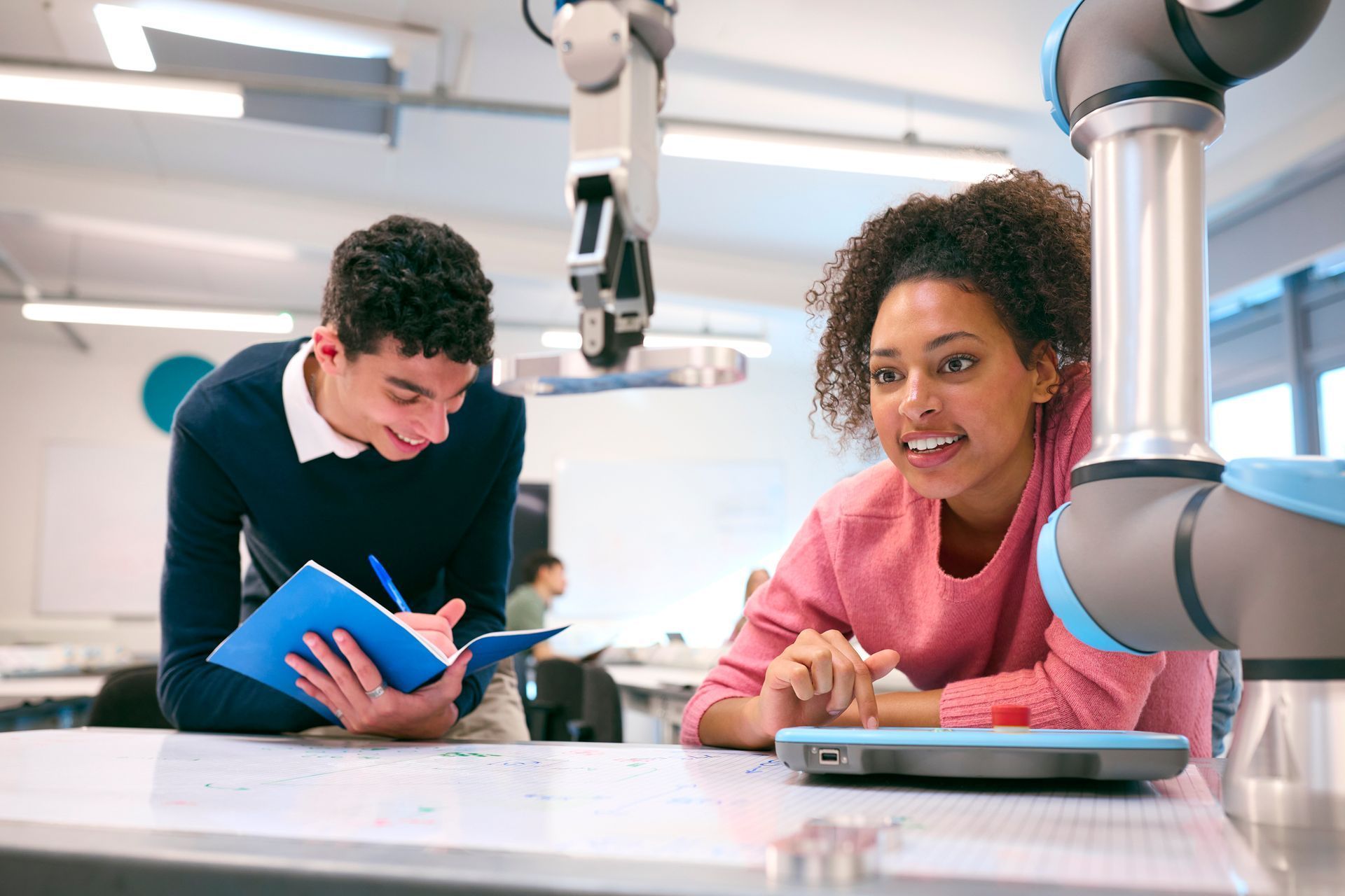 Two students working with a robot arm in a bright classroom, one holding a tablet and smiling.