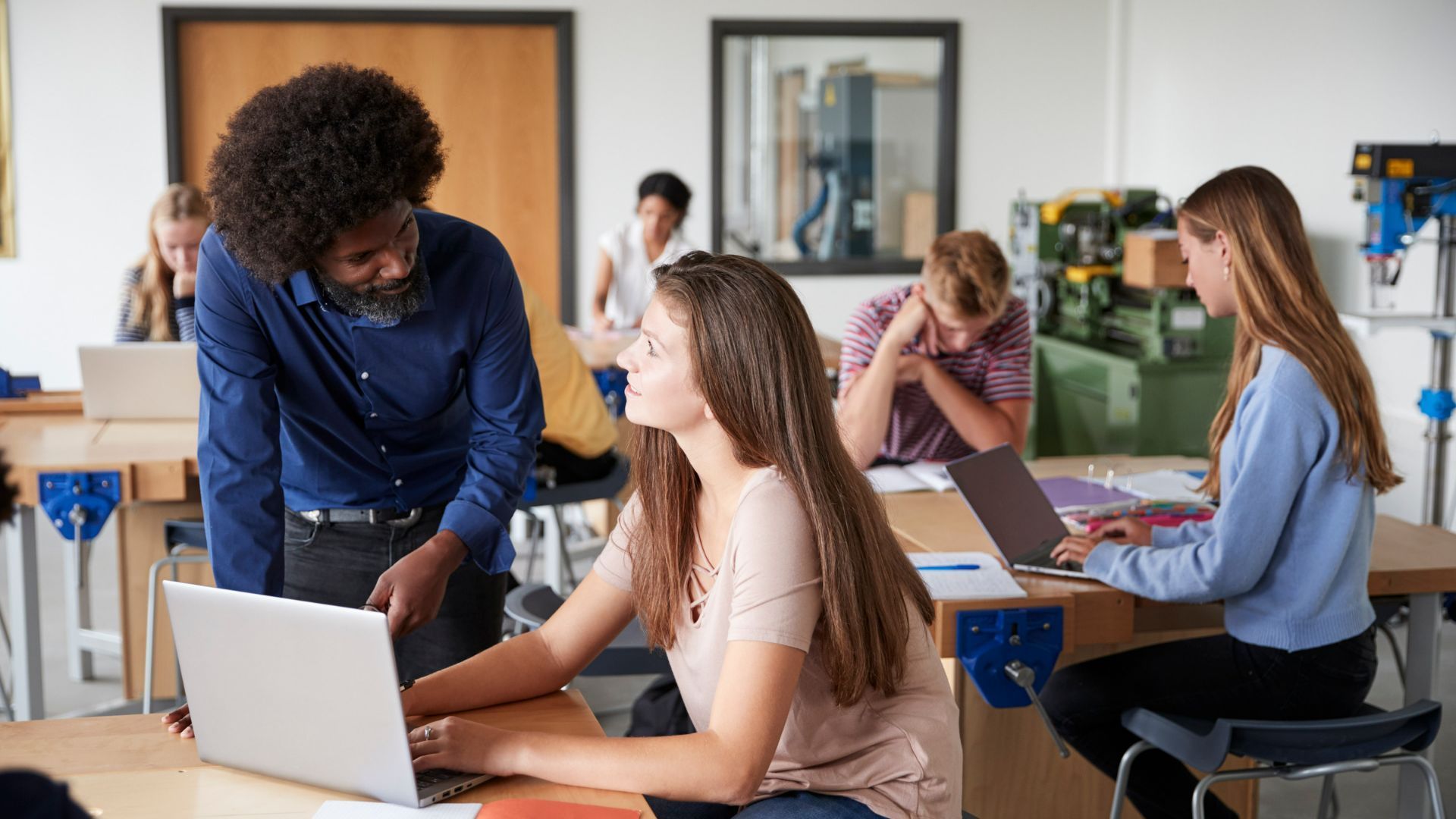 Teacher helping a student in a classroom while others work on laptops.