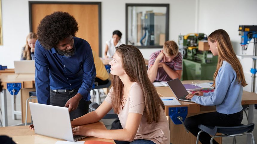 Student and teacher collaborating in a classroom, with laptops and others working in the background