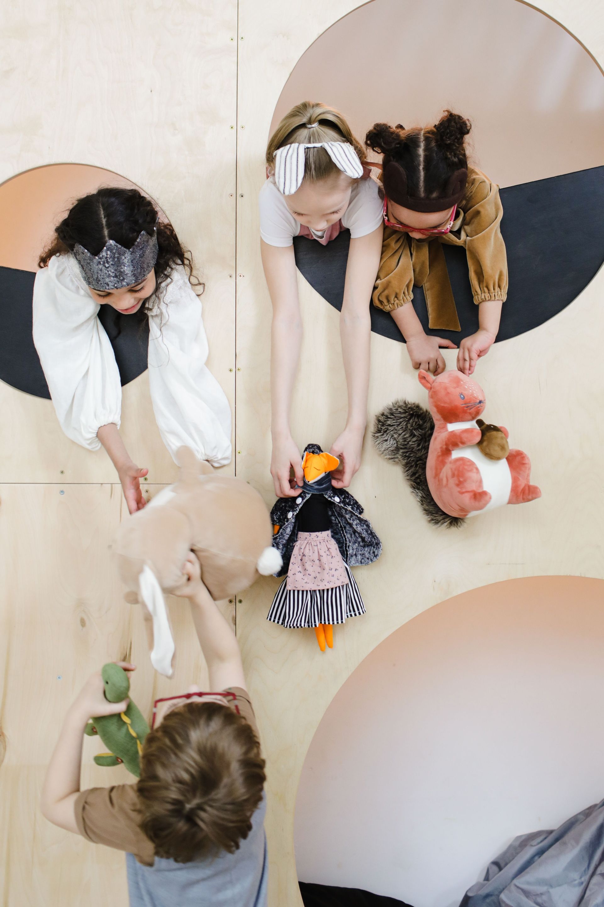 Children playing on a light floor with stuffed animals, viewed from above through round cutouts.