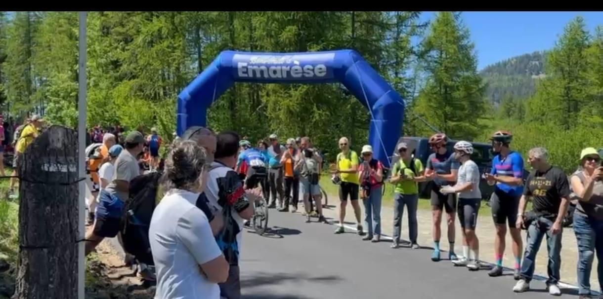 I corridori si sono radunati presso un arco di partenza gonfiabile blu su una strada soleggiata, vicino ad alberi e colline.
