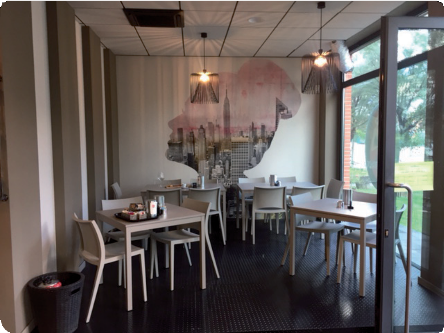 Piccola zona pranzo in un caffè con sedie e tavoli bianchi, lampade a sospensione e un grande murale vicino a una finestra.