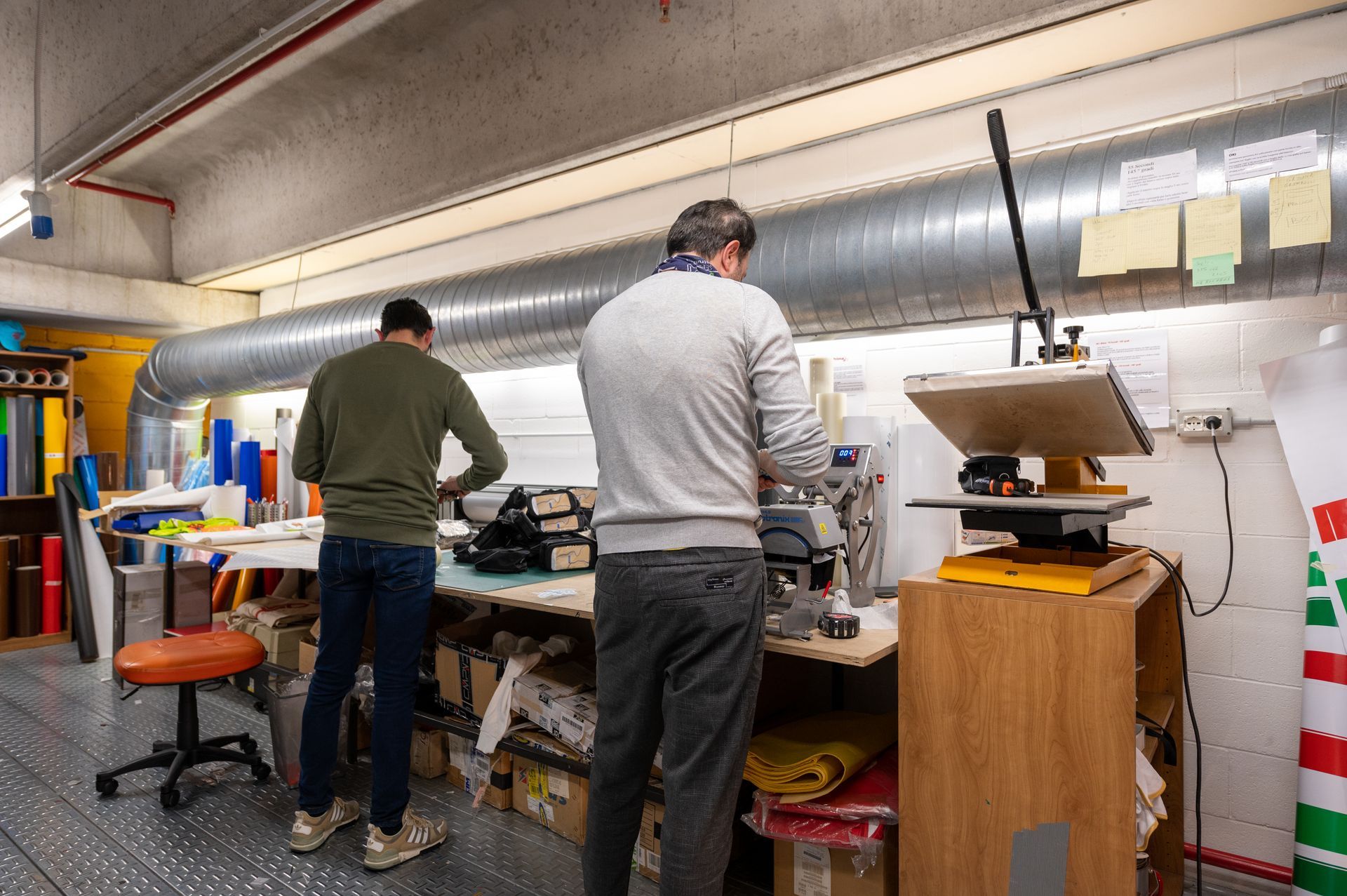 Due persone lavorano a un banco di riparazione ingombro di attrezzi in un'officina, con un grande condotto sopra la testa.