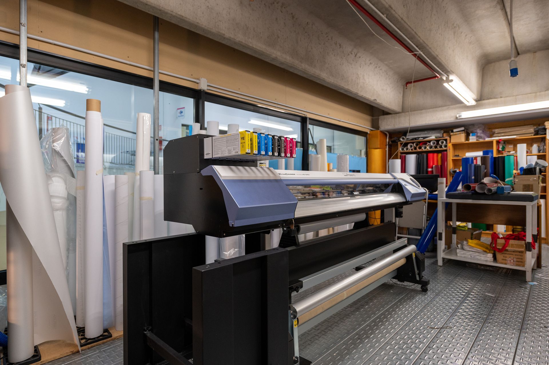 Stampante di grande formato in un'officina, con rotoli di carta e materiali colorati sugli scaffali.