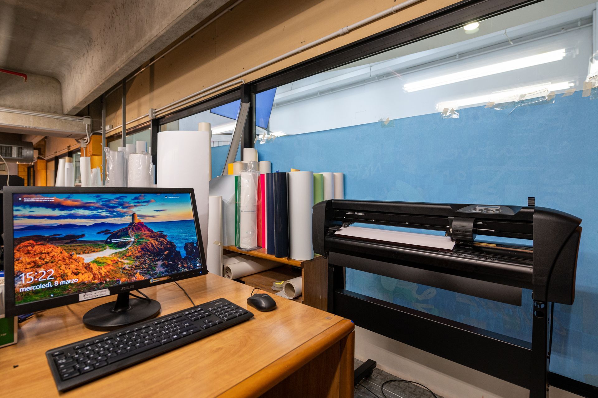 Scrivania con computer e tastiera accanto a una stampante di grande formato in un'officina o in un ufficio, vicino a una finestra.