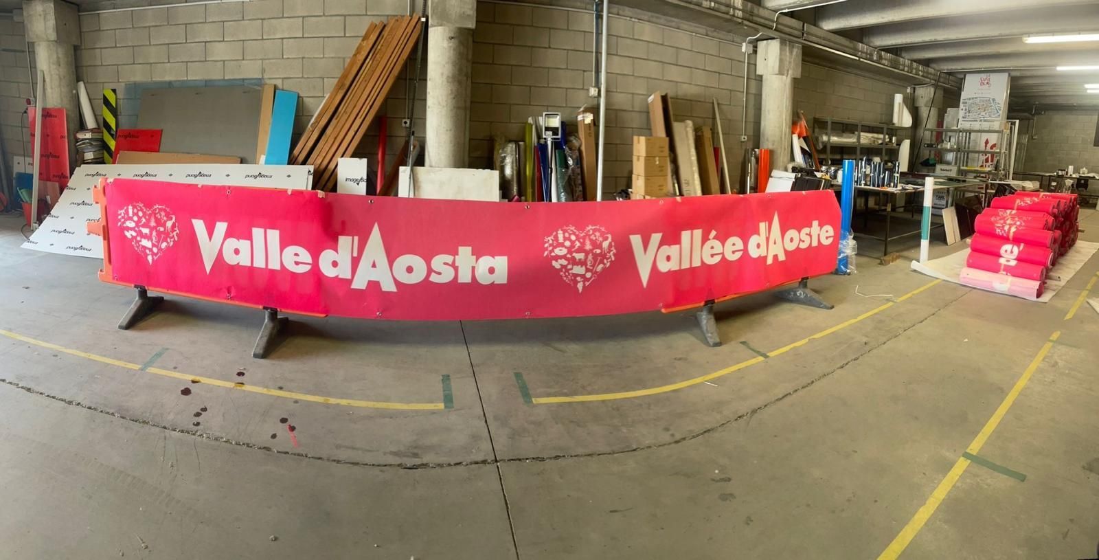 Uno striscione rosso della Valle d'Aosta sovrasta un'officina al coperto, con attrezzi e attrezzature sullo sfondo.
