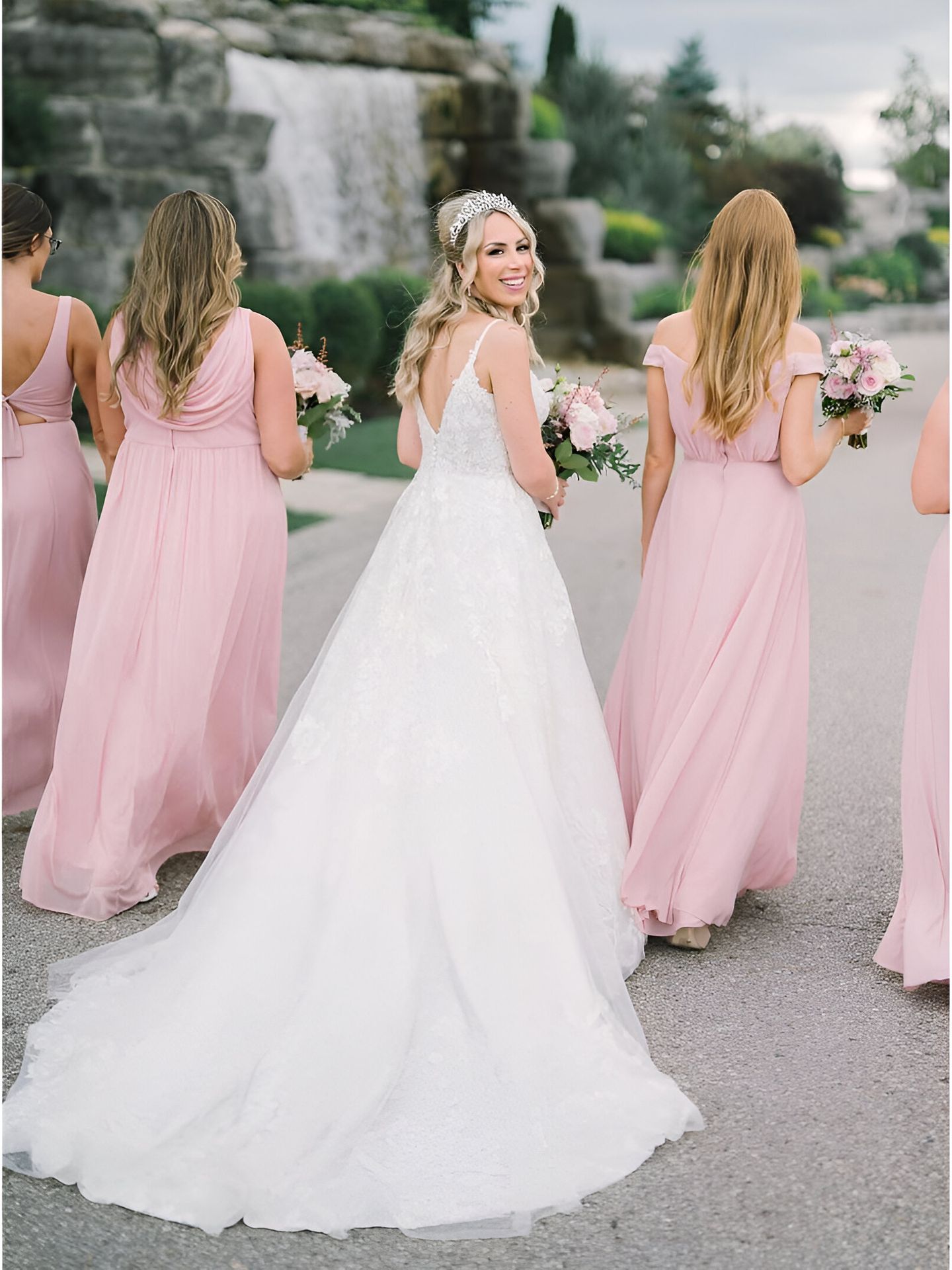Bride in white gown walks with bridesmaids in pink dresses, carrying bouquets.