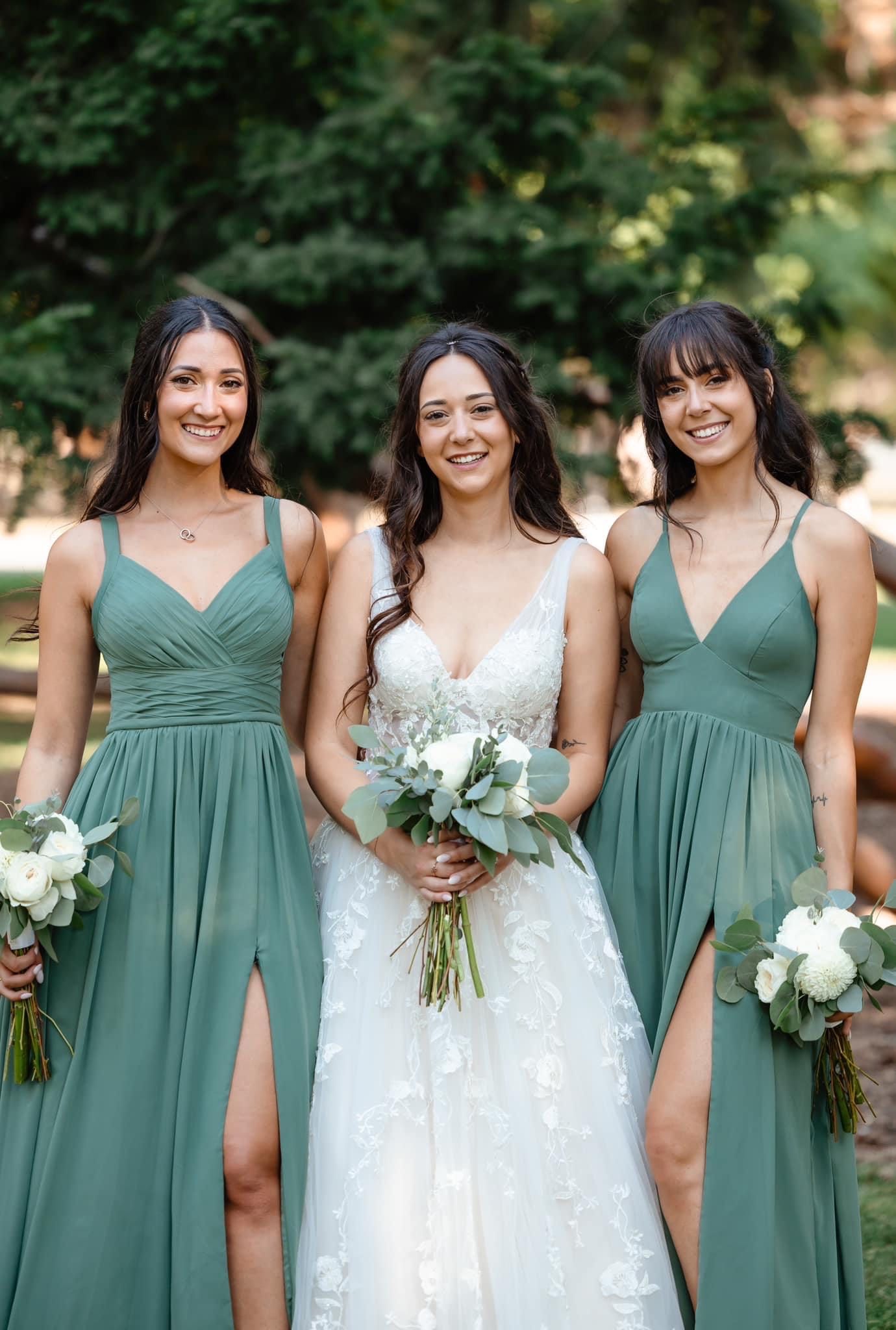 Bride with two bridesmaids holding bouquets, wearing green dresses, smiling outdoors.