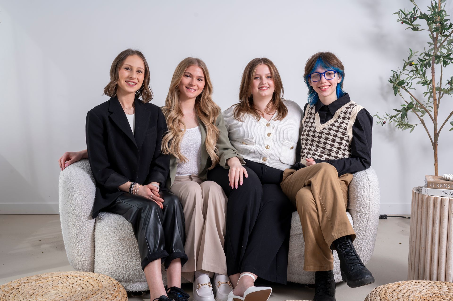Four young people sit on a cream-colored couch, smiling. One wears a black blazer, another light-colored pants. Bright white background.