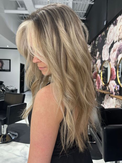 Woman with long, layered, blonde-highlighted hair in a salon; blurred background with floral wallpaper and mirrors.