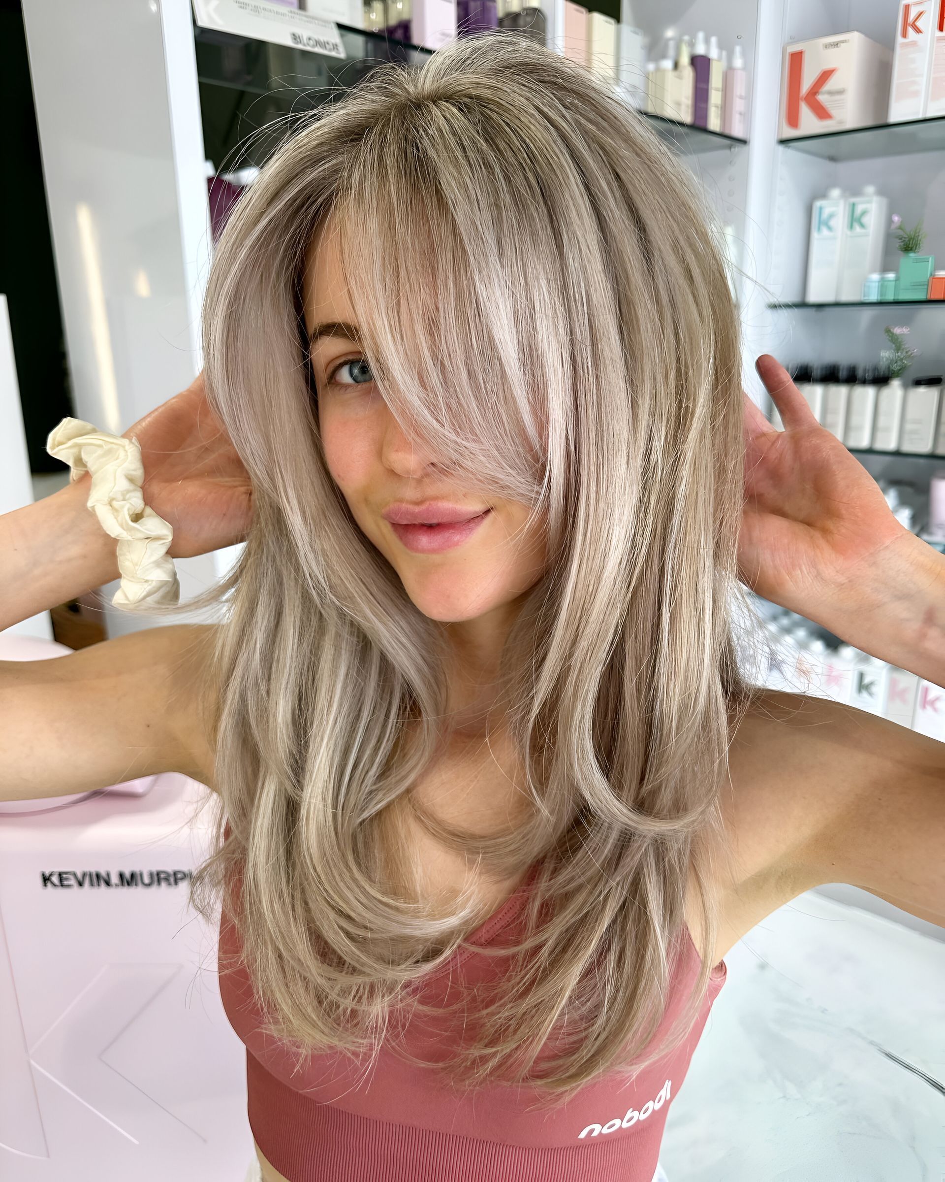 Woman with ashy blonde layered hair, hands framing her face. Pink top, cream scrunchie. Salon setting with products.