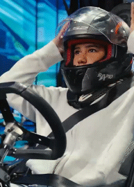 A young girl in a racing helmet and white suit driving a go-kart, adjusting the helmet visor.
