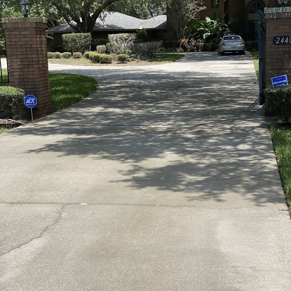 Driveway leading to a house, shadows from trees across the concrete. Brick pillars flank entrance.